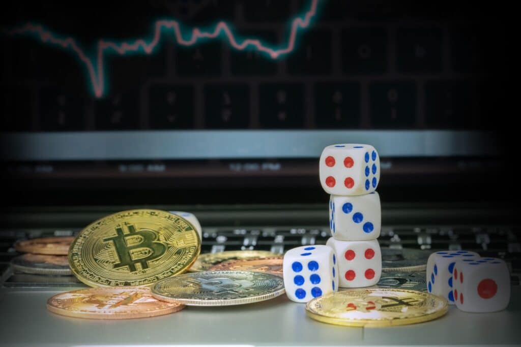 Ein Stapel Würfel und verstreute Bitcoin-Münzen liegen auf einer Tastatur und erinnern an Online-Casinos. Im Hintergrund wird auf einem Computerbildschirm ein Finanzdiagramm angezeigt.