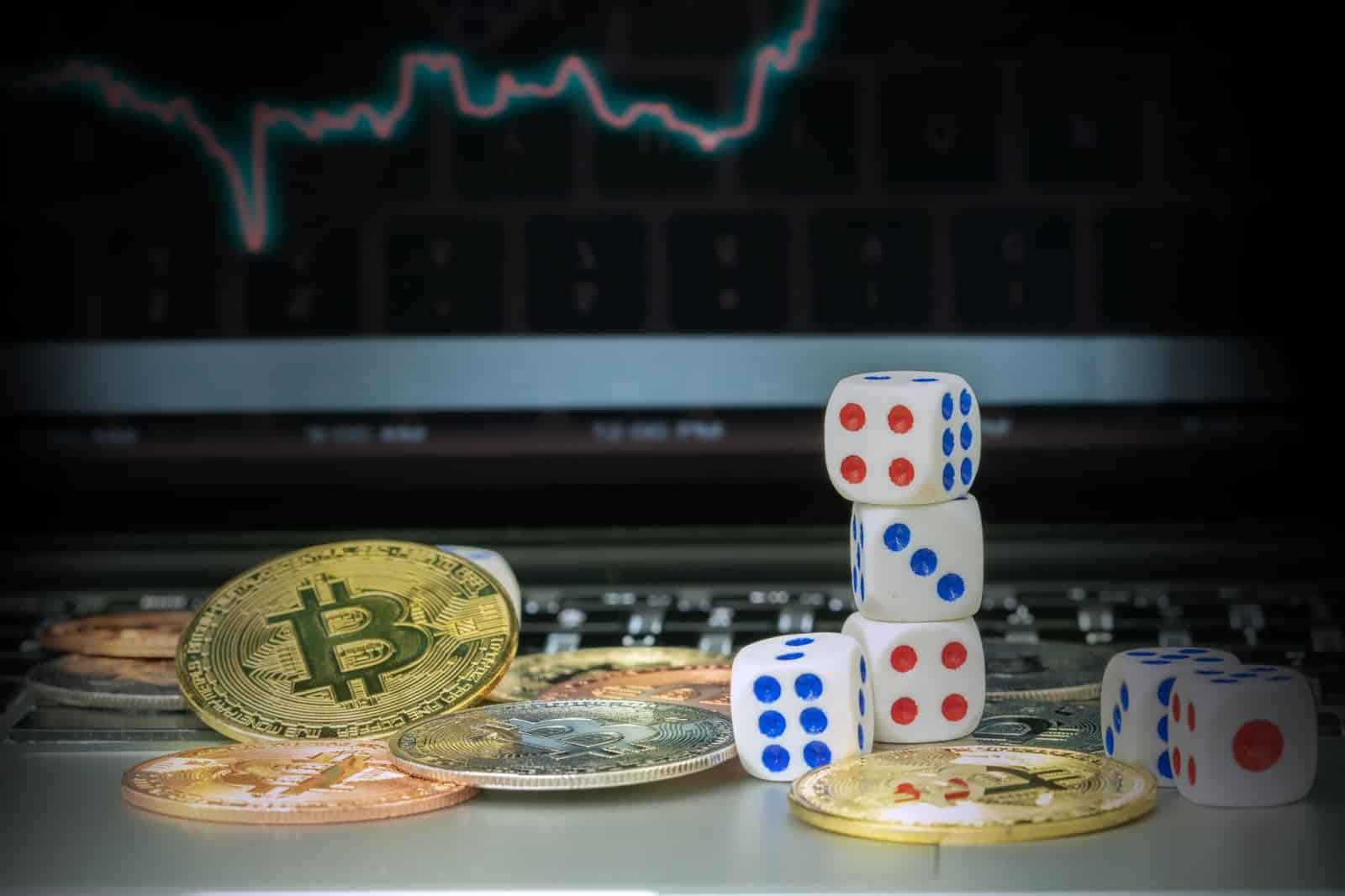 Ein Stapel Würfel und verstreute Bitcoin-Münzen liegen auf einer Tastatur und erinnern an Online-Casinos. Im Hintergrund wird auf einem Computerbildschirm ein Finanzdiagramm angezeigt.