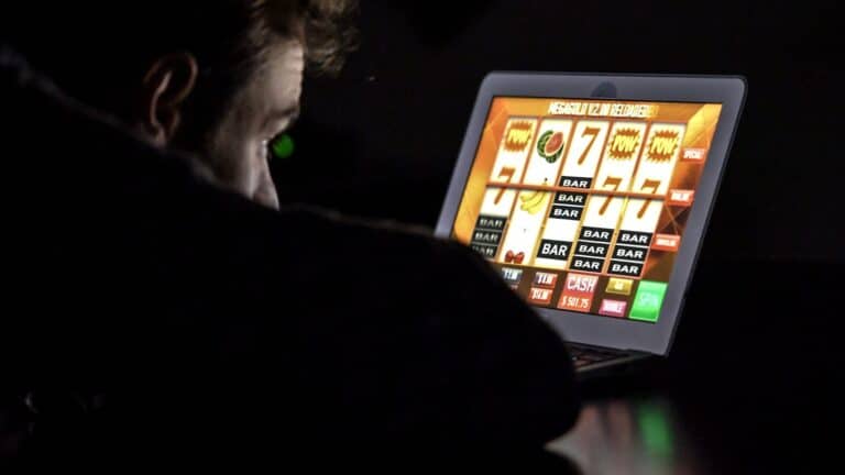 Eine Person sitzt vor einem Laptop, auf dem ein Online-Spielautomat mit verschiedenen Symbolen und dem Wort „SPIN“ angezeigt wird, und recherchiert nach einem Leitfaden zum Steuern auf Online-Casino-Gewinne. Die Szene ist schwach beleuchtet.